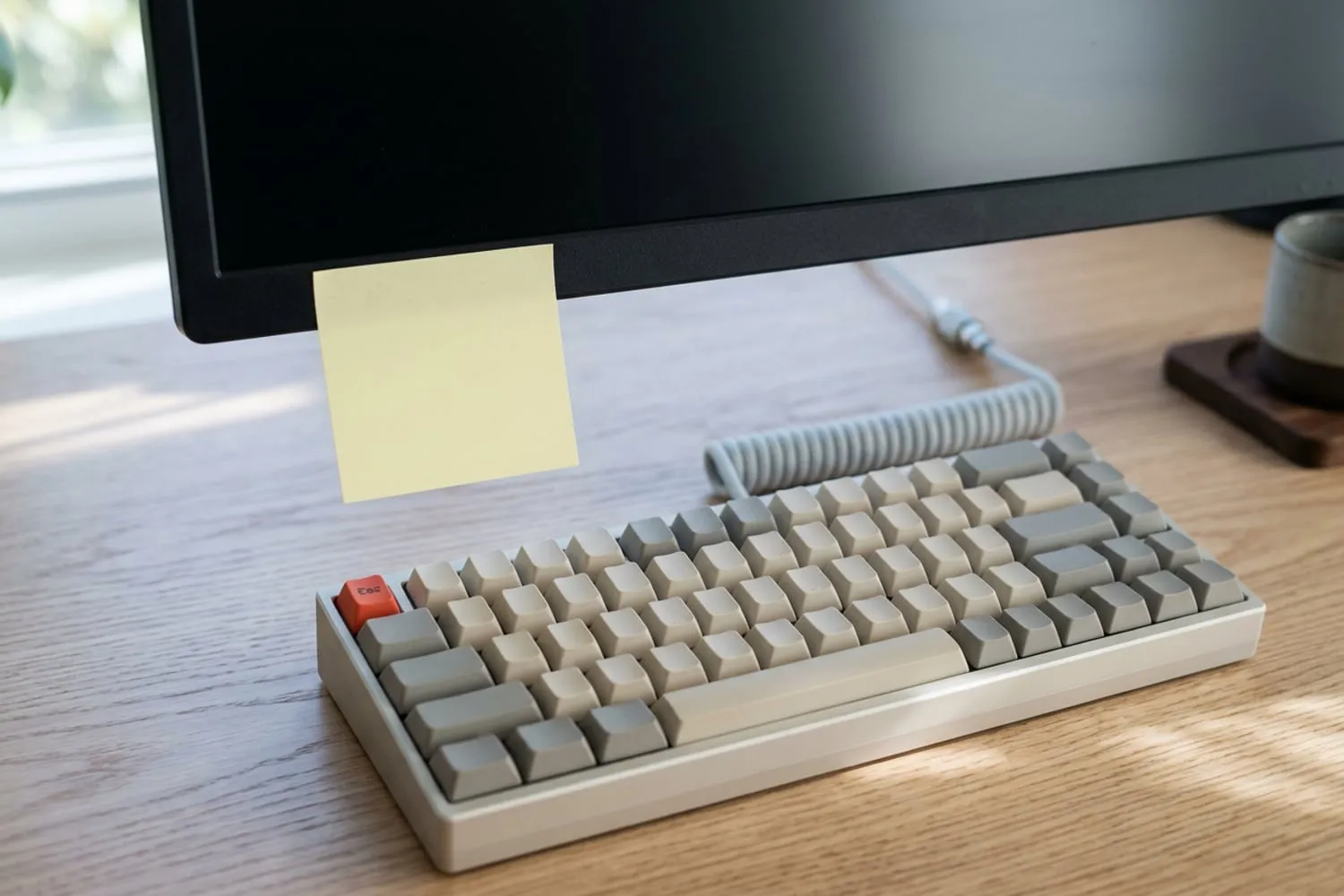 A blank pale sticky note on the lower edge of a dark, switched-off monitor, a compact beige keyboard below it on a light oak desk, every keycap blank