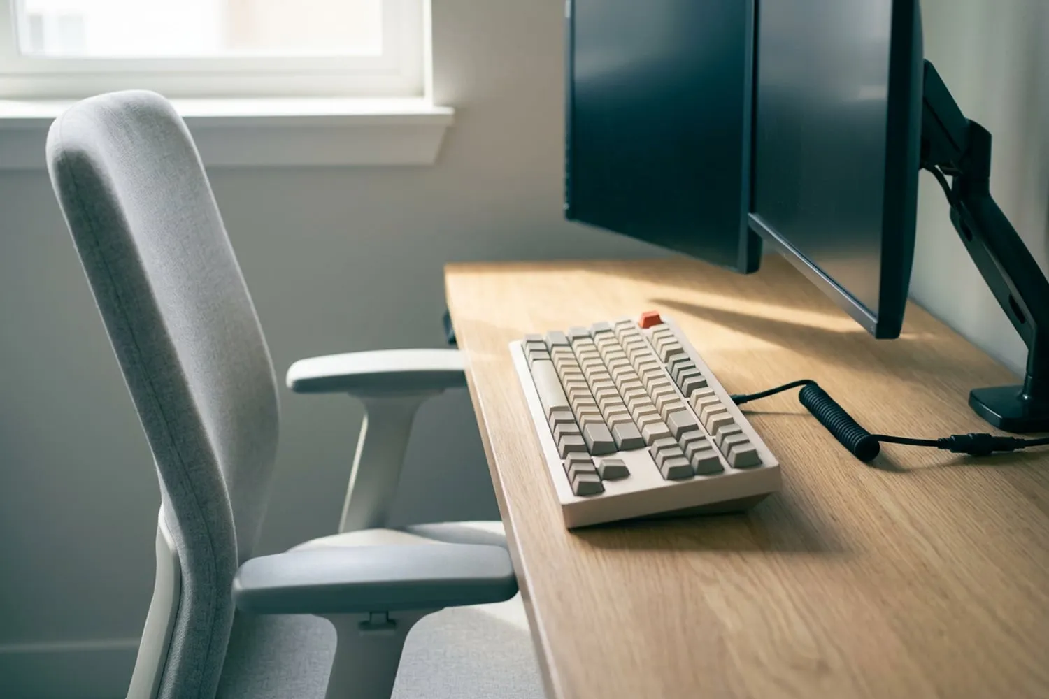 A desk seen side-on in soft daylight: an empty office chair pulled in, a keyboard on the desk at about the height of its armrests, a monitor on an arm behind it with a dark blank screen