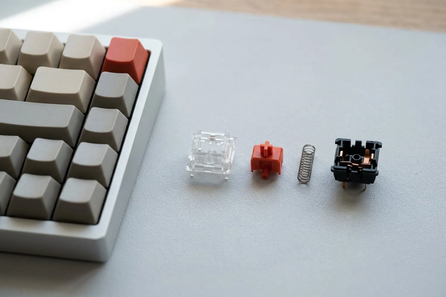A mechanical key switch taken apart on a pale grey desk: the clear top housing, the red cross-shaped stem, the coiled spring and the bottom housing with its two metal pins, all unmarked