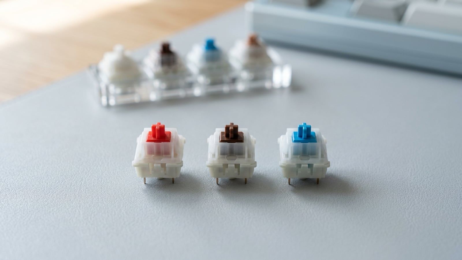 Three mechanical key switches standing in a row on a pale grey desk, one with a red stem, one brown and one blue, their housings unmarked, a small switch tester out of focus behind