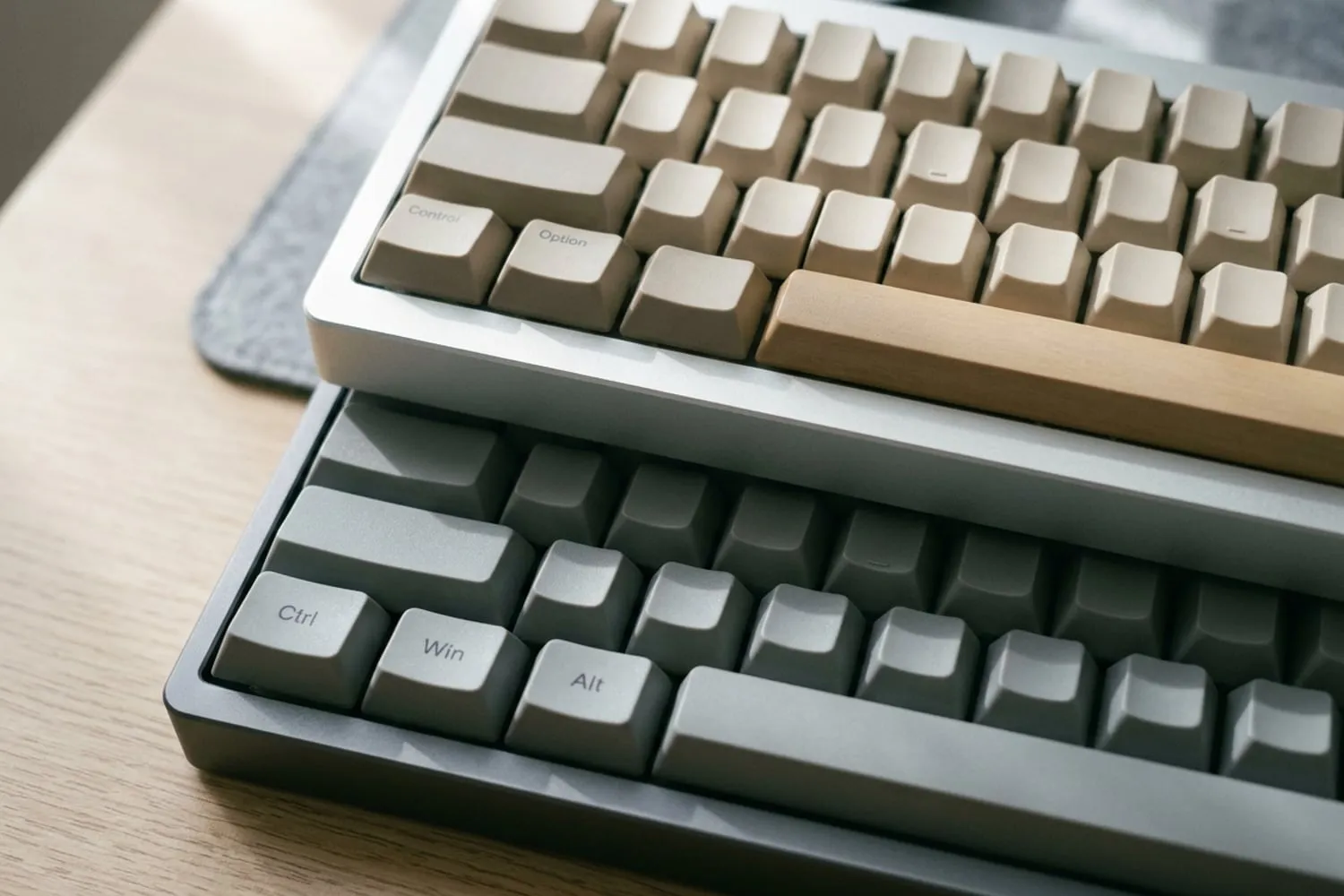 The bottom-left corners of two keyboards close up on a desk, a slim silver one above a dark grey one, their rows of wide modifier keys beside the space bars lined up, every keycap blank