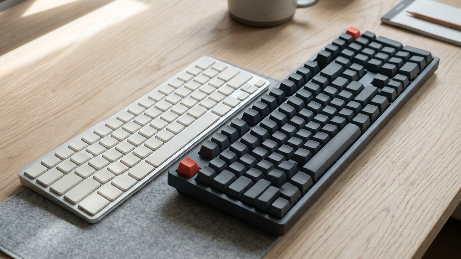 Two keyboards side by side on a pale oak desk seen from above, a slim silver one and a dark grey one with a number pad, their left edges lined up, every keycap blank