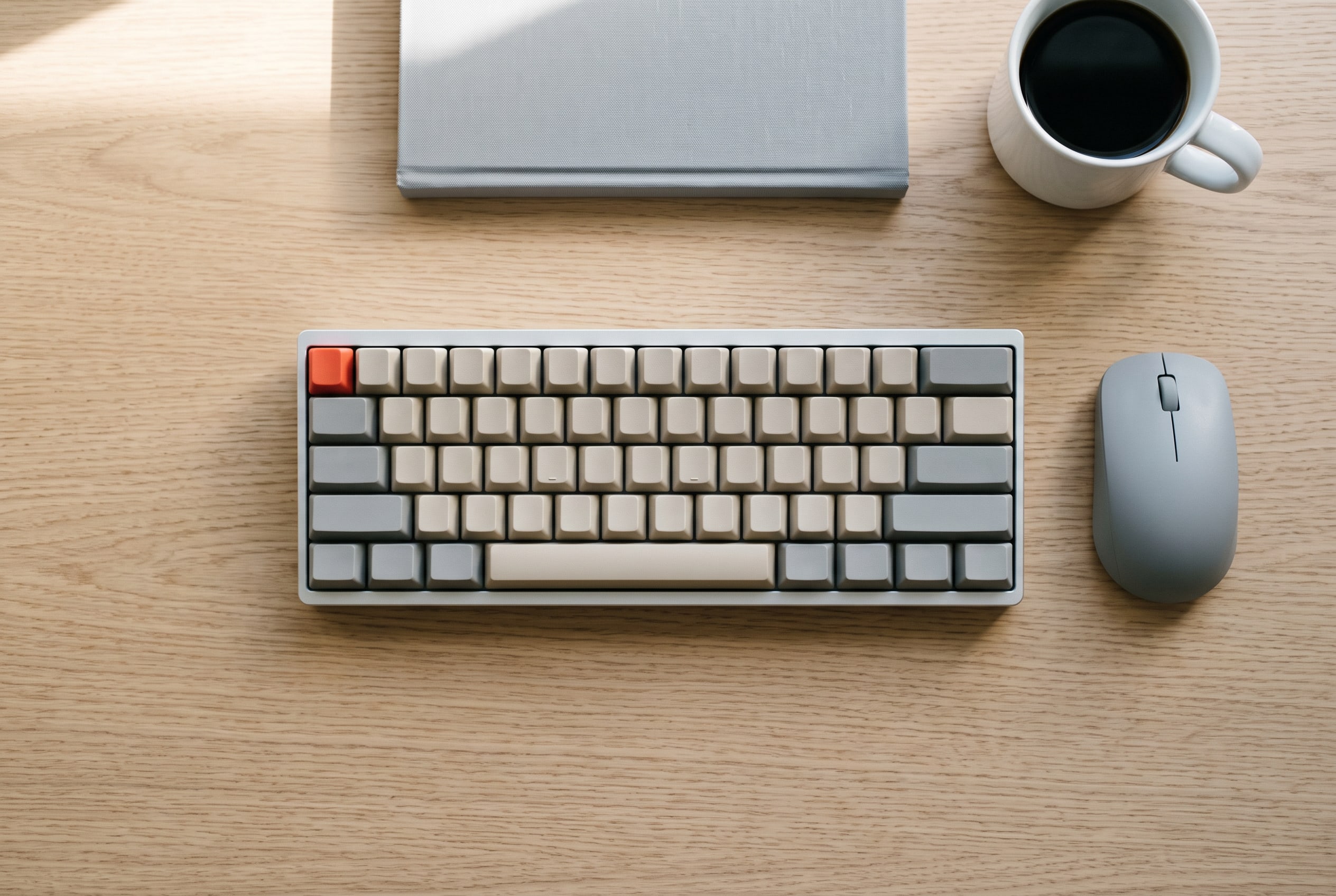 A tidy working desk from above: a beige and grey keyboard with one red key, a grey mouse to its right, a closed grey notebook and a mug of black coffee on light oak.